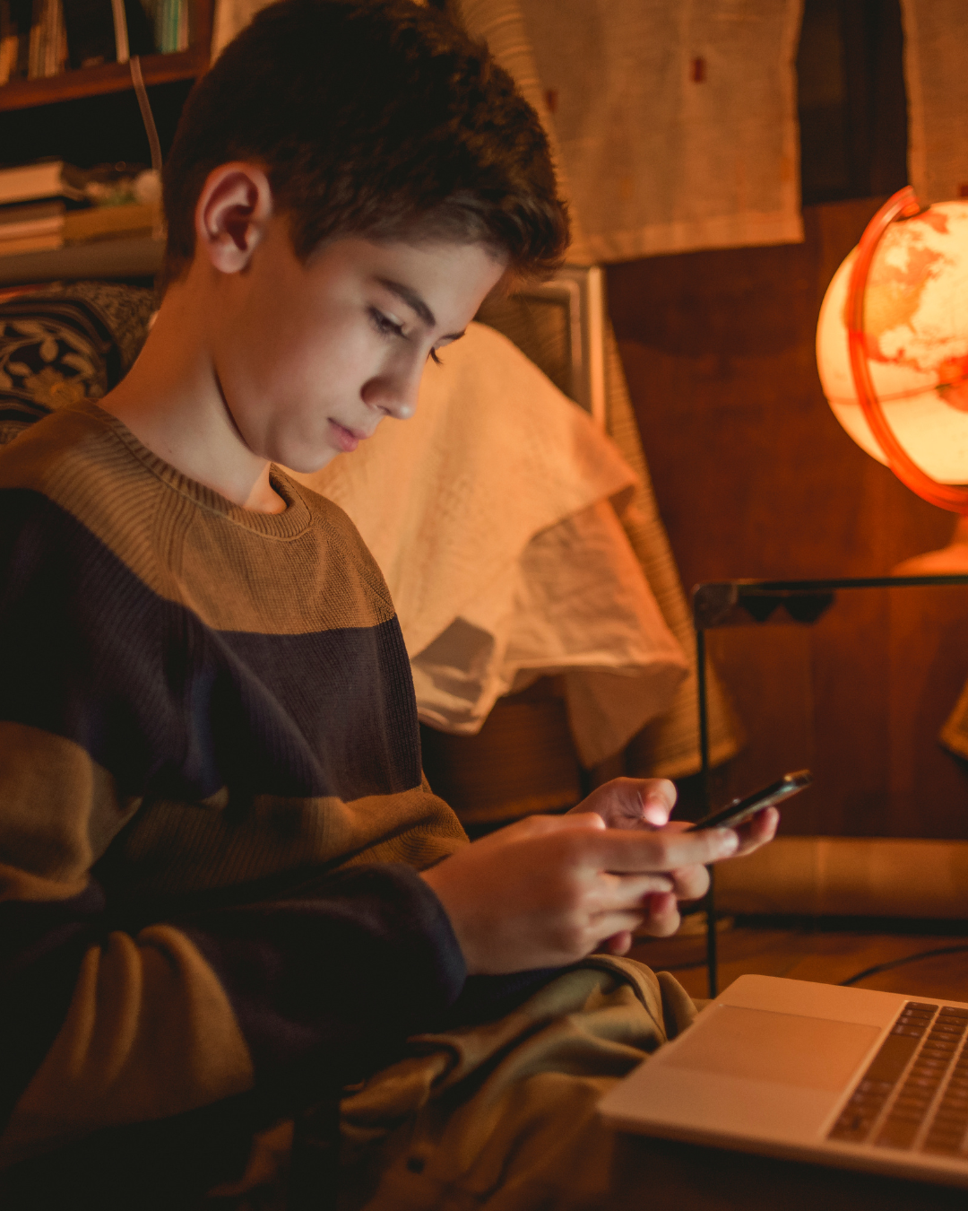 A teenager looking at a phone while using a laptop at home